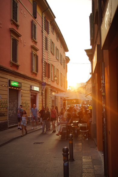 Die Via Delle Moline in Bologna. Die gelbliche, schräg stehende Sonne scheint auf die Gasse, die orangenen Häuser und die Restaurantgäste