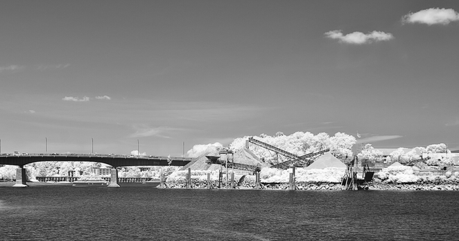 Calm harbour waters. Far shore piles of gravel and conveyor machinery are seen. A busy automobile bridge passes traffic behind the piles.