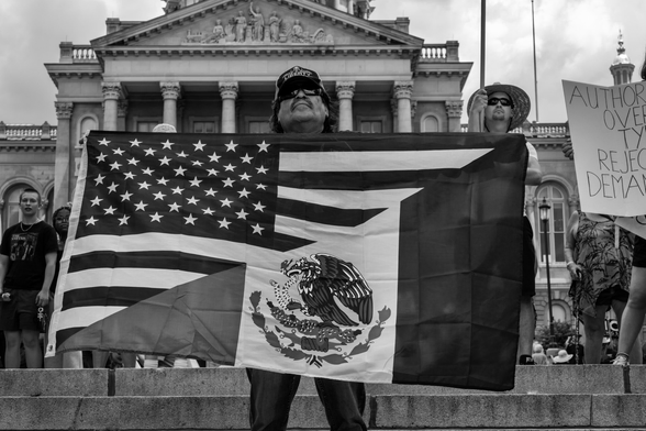 Black and white image of a man in sunglasses holding a flag that’s half US and half Mexican on the steps of the Iowa State Capitol during the No Kings Rally in Des Moines, Iowa. 