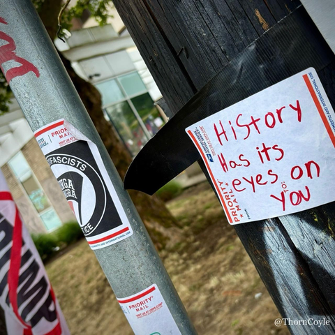 A note scrawled in red ink on a postal label taped to a wood utility pole reads “History has its eyes on you.” 

A metal pole next to it has an anti fascist sticker. A building is seen between the two poles.