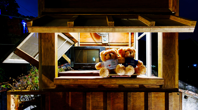 Art installation with two teddies wearing “I Love Taipei Media School” t-shirts sitting on the window sill of a make-believe apartment with open windows and a fan and a tv-set in the background.