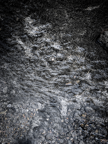 A dark, high-contrast, top-down shot of water flowing over a rocky, uneven surface. The water itself shows ripples, small waves, and streaks of light.