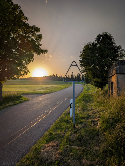 A serene rural scene at sunset. A paved road curves gently to the right, disappearing into the distance. On the left side of the road, a lush green field stretches out, with the sun's rays visible peeking over the horizon, casting a golden glow. The sun itself is partially obscured by the distant tree line, creating a beautiful sunburst effect. On the right side of the road, tall, wild grasses grow alongside a somewhat dilapidated, concrete structure. In the foreground, an empty road sign with a distinctive triangular shape on top stands beside the road. 