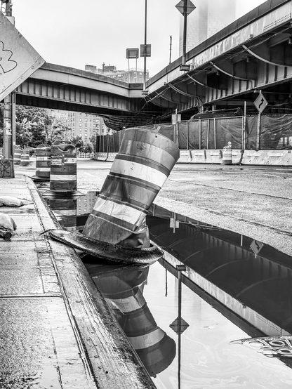 A battered construction barrel on the side of a city street. The barrel is reflected in a puddle 