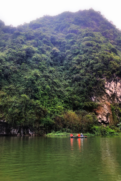 A limestone mountain covered in green vegetation, with a bare cliff face at right frame, rises over the calm waters of a wide river. At the far edge where the river meets the mountain is a rowboat with passengers wearing red life jackets that are reflected in the water. The rower wears a conical hat. The scene contrasts the smallness of the human figures with the expansive scale of their surroundings.