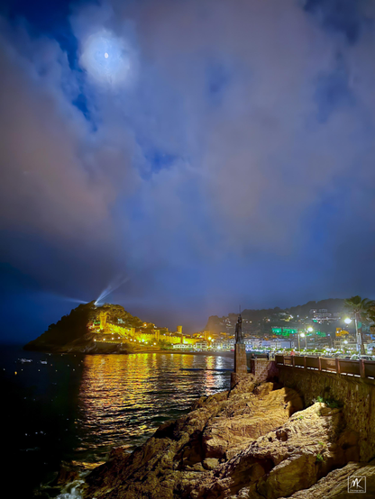Nighttime color photo of a waxing moon shining through low clouds above a coastal city.