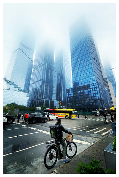 A deliverista on his bike waits by the sidewalk at a wide city intersection. Pedestrians weave between cars on the crosswalk stripes. A red and yellow tour bus inches into the intersection to make a turn. Blue-toned glass skyscrapers, with grids of reflective windows, rise up and disappear into the fog.