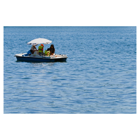 A small white and blue pedalo boat floats on calm blue water. Three people are seated on the boat under a white umbrella for shade. One person appears to be wearing bright green or yellow clothing. The boat is surrounded by open water that extends to the horizon.