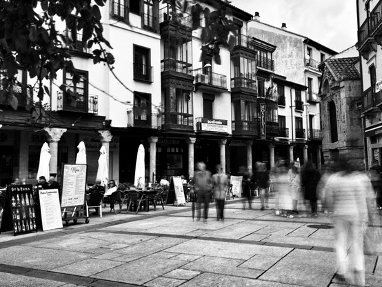 Foto en blanco y negro de una plaza con edificios antiguos de varios pisos con balcones.  Las plantas bajas de los edificios albergan negocios, algunos con mesas al aire libre.  Se ven personas, borrosas por el movimiento, caminando por la plaza.  Hay sombrillas y carteles de menú visibles en las terrazas de los cafés. La atmósfera es tranquila y urbana.