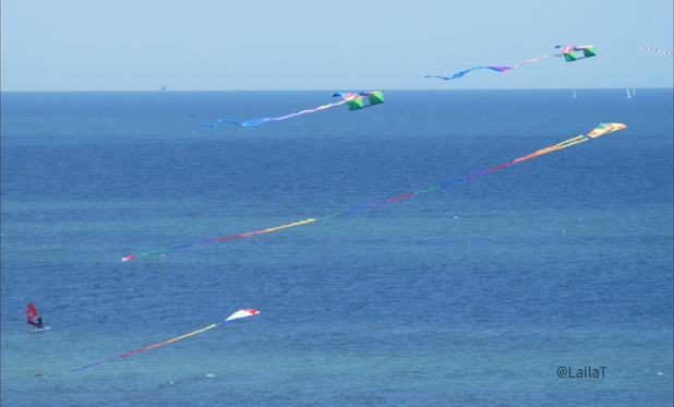 Vier bunte Drachen schweben über dem Meer, das sich in Blau- und Türkistönen zeigt. Auf dem Wasser sind mehrere Segelboote, ein Surfbrett und ein Schiff zu erkennen.