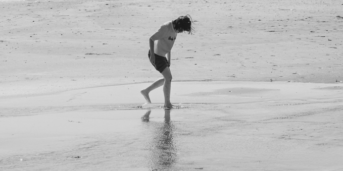 A person with long hair walks barefoot on a wet, sandy beach. The individual wears a light-colored shirt and dark shorts, casting a faint reflection on the sand. The background features a vast expanse of flat, smooth sand extending to the horizon. The image is in black and white, emphasizing contrasts and details.