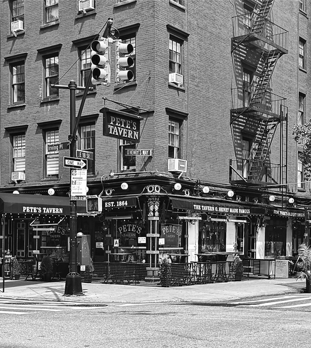 Black and white photo taken from the perspective diagonally across the street from a corner. It is an old pre-war brick building with a sign that says Petes Tavern, and awning that says Petes Tavern established 1864. The awnings are black and there are twinkle lights around them. There are also large round lights approximately 5 feet apart going across the upper crown molding before the brick starts. On the right side of the brick building are fire escapes with the shadows reflecting onto the wall. In the foreground can be seen some of the white stripes of crosswalks 