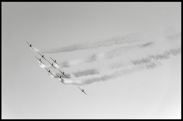 A photo of nine jets belonging to the Canadian Forces Snowbirds fly in formation during a Fleet Week demonstration in the skies above San Francisco. Smoke trails off from the tails of most of the jets as they arc through the sky. 

Minimalist, monochrome, magical.