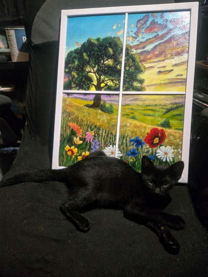 A black kitten lying in front of a colorful painting of a wildflower meadow seen through a window.