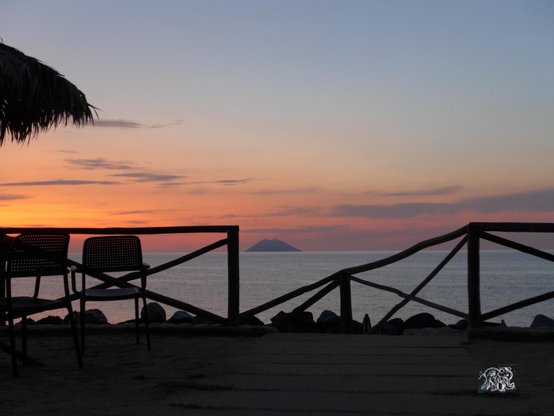 Vorne  dunkler Strandschirm, Geländer und Sessel, im Hintergrund Meer mit Sonnenuntergang und Stromboli