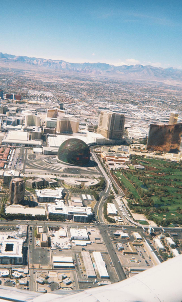 This is a daytime image of Las Vegas taken from the window of a commercial passenger airplane as it takes off toward the northwest from the Harry Reid Airport (formerly McCarran Airport) In the center of the photo is the now iconic Sphere, which from the air, looks like a a bubble sitting on the ground. A green golf course, multiple industrial buildings and several hotel/casino buildings are in view. In the distance, the mountains that surround the greater Las Vegas valley are in view below a clear blue sky. The photo was taken using an Apple iPhone 16 pro plus.