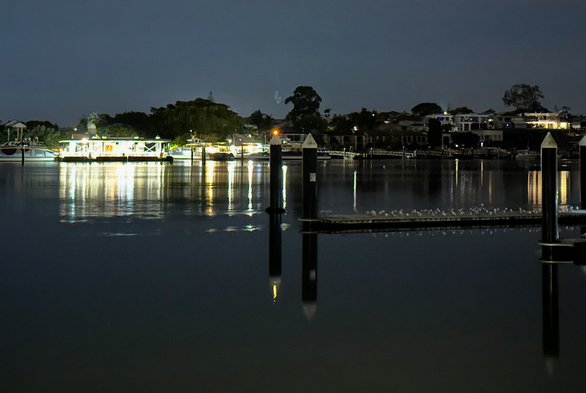 Looking across the Brisbane River at night. On the opposite bank, the ferry terminal is lit up in a blaze of light, reflecting on the calm river surface. Along the bank a few other lights are on. In the foreground, close to the near bank is an empty dock, its piers reflected in the water. A large flock of seagulls is roosting on the empty dock.