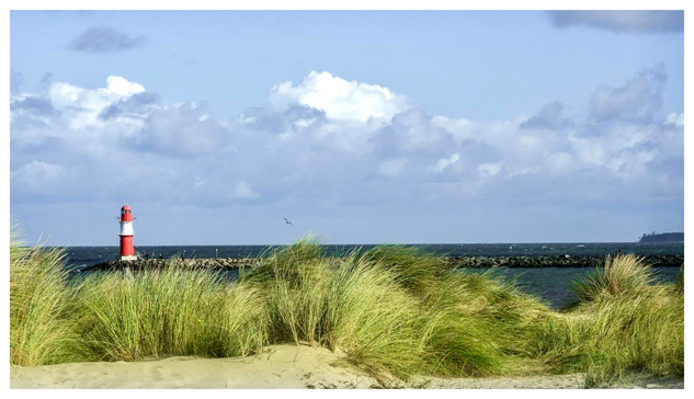 A scenic view featuring a red and white lighthouse on a rocky pier in Warnemünde with sandy dunes and tall grasses in the foreground. The background shows the calm Baltic Sea under a partly cloudy sky.