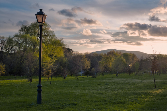 Fotografia de un prado de hierba verde en el margen de un rio, donde en primer plano a la izquierda se ve una farola, tras ella se ve una hilera de árboles que delimitan la orilla del río, tras estos se vislumbra la linea de unas montañas en la lejania, en la parte superior un cielo con nubes que en el lado derecho esta iluminado intensamente por la luz del sol al atardecer