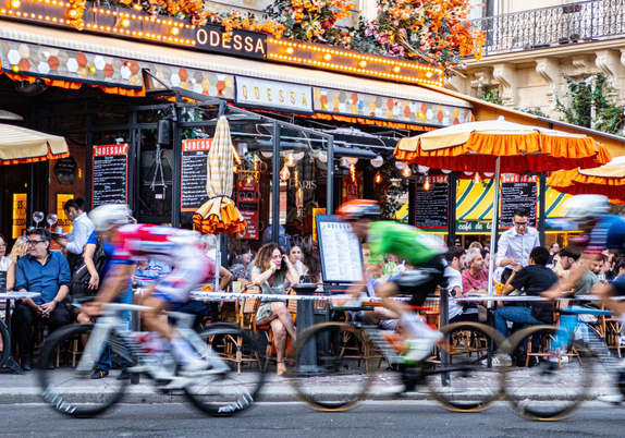 A picture of a Parisian café called Odessa with orange flowers and trimmings, seen from across the road, with a number of cyclists passing rapidly by in the foreground. The customers are being served and there is a woman in the centre talking on the phone. The cyclists have been blurred by using a relatively slow shutter speed.