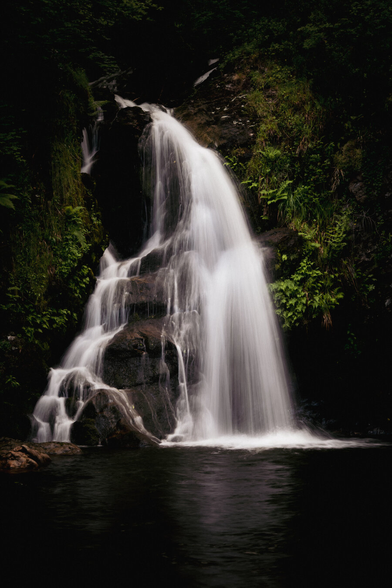 a small waterfall surrounded by greenery, with a subtle reflection in the river that it feeds. Some vignetting and darkening has been added to make the image feel a little moody