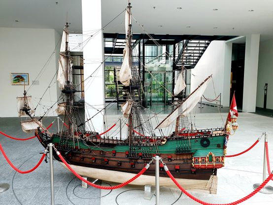 Large model sailing ship with green hull and white sails displayed in a modern museum gallery. The detailed replica features multiple masts with rigging and decorative stern elements. Red rope barriers on metal posts surround the exhibit. Modern architecture visible in background includes stairs, large windows, and white walls with contemporary lighting.