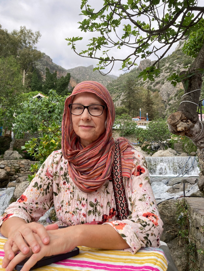 A lady sitting at a table in the city of Chefchaouen. Behind her many green trees and dry hills can be seen. A waterfall is behind her left shoulder.