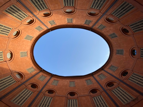 A circular view of the sky framed by an orange, domed structure with evenly spaced round windows and vertical vents. The architectural design creates a striking contrast between the building's warm colors and the blue sky above. Ferrara