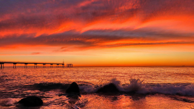 La imatge mostra una posta de sol espectacular sobre l'oceà. El cel està tenyit de tons ataronjats i vermellosos vibrants, creant un efecte dramàtic. A la distància, es veu un pont llarg que s'estén sobre l'aigua. En primer pla, les onades trenquen contra les roques de la riba, reflectint els colors del cel. L'escena transmet una sensació de serenitat i bellesa natural.