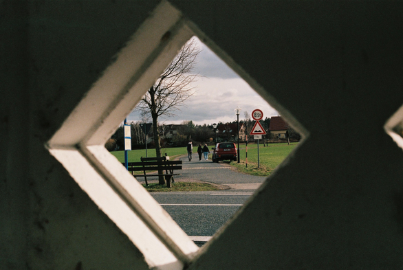 [english below] Das Foto zeigt 3 Menschen, welche eine Straße entlang laufen. Die Menschen laufen in Richtung Bildhintergrund wo Häuser stehen. Das Foto wurde durch ein kleines Fenster (ohne Glas) einer älteren Bushaltestelle aufgenommen.

[english] This photo shows three people walking down a street. The people are walking toward the background, where houses are located. The photo was taken through a small window (without glass) of an older bus stop.