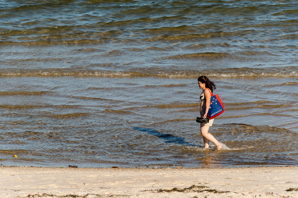 A young woman walking in the water at the beach edge.  Her arm half covers the Australian flag on her bag, which also has the word Australia across the bottom of it.