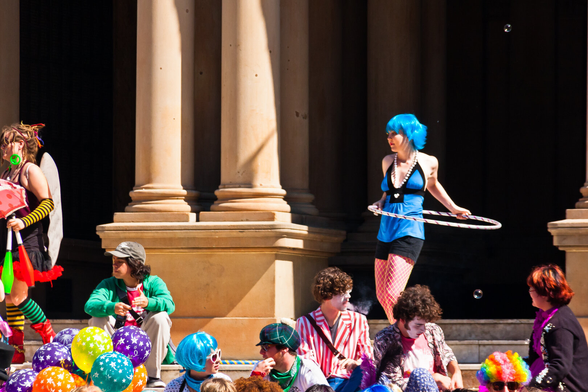 A group of young people in multi-colored clothing and hair or wigs. This looks like it must have something to do with an event of some kind. On the right, a girl has a hula hoop around her. In the background, there are two classic looking  building columns.