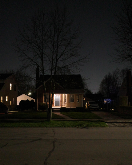 Color photograph looking across a residential suburban street at night at a house with its porch light on, a tree in the front lawn splays out bare branches against a clear silvery night sky