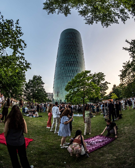 Das Bild zeigt eine Parkszene mit vielen Menschen, die in Köppchen herum stehen und im Vordergrund zwei Frauen, die eine lilane Decke ausbreiten. Dazwischen einige grüne Bäume und im Hintergrund das Frankfurter Hochhaus, dass im Volksmund das Gerippte genannt wird.