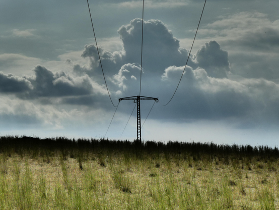 Wir schauen auf einen Strommast inmitten einer steppenartigen Landschaft in Mitteleuropa. Die Stromkabel scheinen das Bild zu zerteilen. Über allem prangt ein monumentaler Wolkenhimmel.