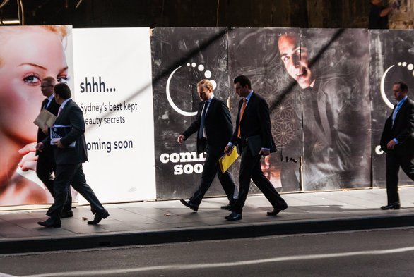 Men in business suits walk past a construction hoarding with a woman's face on the left and the following words on it: Shhh... Sydney's best kept secrets. Coming soon.