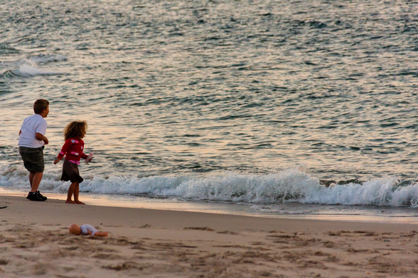 On the left, a boy, and a girl to his right, stand at the waters edge with arms raised. This body language seems to suggest that they are perhaps about to run back from the incoming wave. Behind them, there's an out of focus doll, face down on the sand. The photo background only shows see and sand, so there are no contextual clues as to where this was taken.