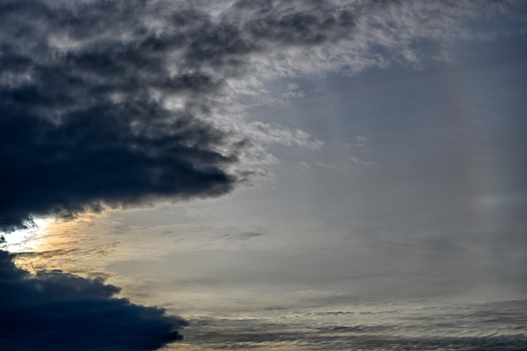 Color photo of clouds in the sky. On the left, they are thick and dark, but an angled gap reveals the bright light of the hidden sun. In the middle and the right, thin wispy clouds are present. A rainbow arc, barely visible, is on the extreme right of the frame.