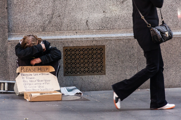 Against a stone wall of a city building, a man is crouching or possibly sitting on something, with his head downwards and face covered by his arm.  By the look of his hands, though, he may be quite young. In front of him, there's a box for collecting money and a sign that says:  Please help. Broken leg and doing it tough. All help very appreciated and put a good use. Thank you and God bless. To his right, a woman with a fashionable handbag, but only visible from shoulder level downwards, walks past.