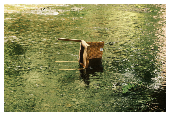 Ein umgekippter Stuhl aus geflochtenem Material steht mitten in einem flachen, grünen Fluss. Zwei Stuhlbeine ragen schräg aus dem Wasser, während die Sitzfläche teilweise untergetaucht ist. Auf der Seitenfläche des Stuhls ist ein kleiner QR-Code sichtbar. Das Wasser fließt ruhig, mit sanften Wellen und Lichtreflexionen, und im Vordergrund treibt ein kleiner Ast mit grünen Blättern. Die Szene wirkt surreal, fast wie eine stille Installation in der Natur.