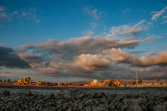 This image shows a construction site at sunset. Heavy machinery is visible on a dirt field. The sky is dramatic, with clouds illuminated by golden light. Mountains can be seen in the background. Yellow construction vehicles, including a bulldozer and a grader, are positioned on the site. The foreground displays rough, uneven soil.