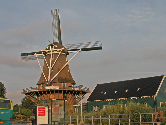 Photo by Fenichel

A windmill in Holland is on the left side, ;with its brick bottom, black and white blades, 3 windows at the top, and blue sky hosting its winds. On the right is a green building with sloped roof, and in the foreground/bottom is a fence along the road.