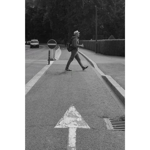 Black and white street photography taken in Luxembourg city. A man with a hat is crossing a bike lane with a can of soda in his hand. His legs are just above the end of an arrow sign painted on the ground.