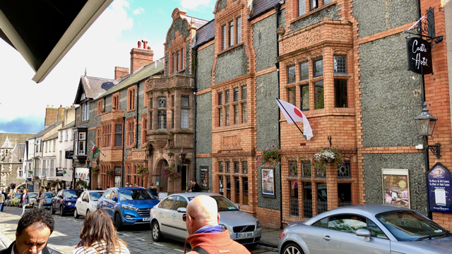 This picture captures a charming street scene in what appears to be a historic town. The buildings are characterized by traditional architecture with a mix of red brick, stone, and ornate detailing. The prominent structure on the right side is identified as the "Castle Hotel," with its sign hanging on a black iron bracket and a Japanese flag displayed outside. The hotel features large bay windows and hanging flower baskets, adding to its quaint appearance.

The street is lively with pedestrians walking along the cobblestone pavement and several parked cars lining the road. Other buildings in the background showcase similar architectural styles, with flags, including the Welsh flag, displayed. The sky is partly cloudy, allowing some sunlight to illuminate the scene, enhancing the vibrant and inviting atmosphere of the town.