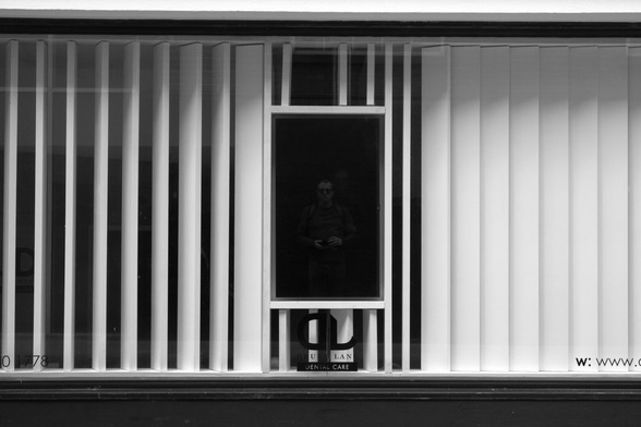 Monochrome shot of a wide commercial window, across which are large vertical blinds, half-open on the left, closed on the right. In the middle is a portrait oriented monitor, unpowered and black, in which the photographer can just about be seen.