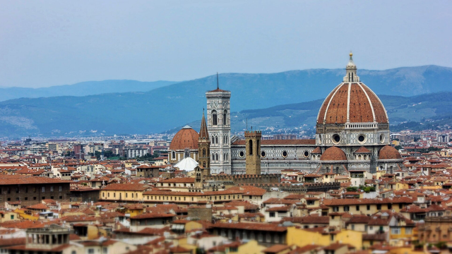 An elevated, slightly blurred view of the terracotta rooftops of Florence, Italy, with the prominent Florence Cathedral (Duomo) and its large red-tiled dome and Giotto's Bell Tower dominating the centre. In the background, blue mountains rise against a light grey sky, and the cityscape stretches out towards the horizon.