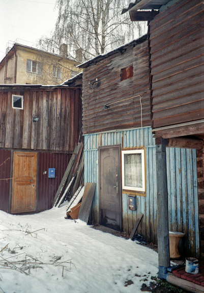 The photo shows an old wooden house with a porch. Part of the house is dark, part is painted blue. There is snow at the house.