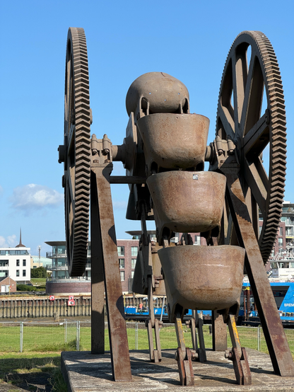 Eine große, verrostete Industriemaschine mit großen Zahnrädern und mehreren vertikal gestapelten schalenartigen Strukturen erinnert. Im Hintergrund sind moderne Gebäude und ein klarer blauer Himmel zu sehen.