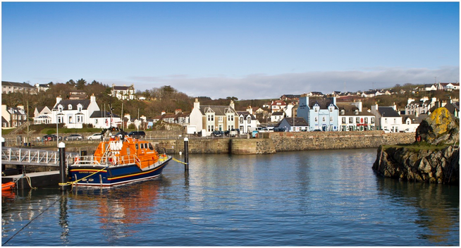 A picturesque harbor scene featuring colorful buildings along the waterfront. An orange RNLI rescue boat is docked, with a stone pier and rocky shoreline in the foreground. Calm water reflects the clear blue sky, creating a serene coastal atmosphere.