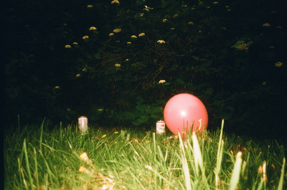 A ground-level photo of two beer cans and a smiling balloon next to the can on the right on grass in front of some flowery bush. The grass in the foreground is very bright because of the camera flash